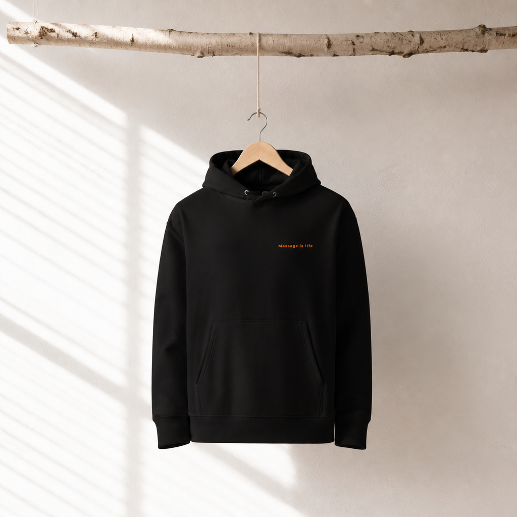 Sweat à capuche noir bio avec inscription 'Massage is life', suspendu à une branche, lumière naturelle