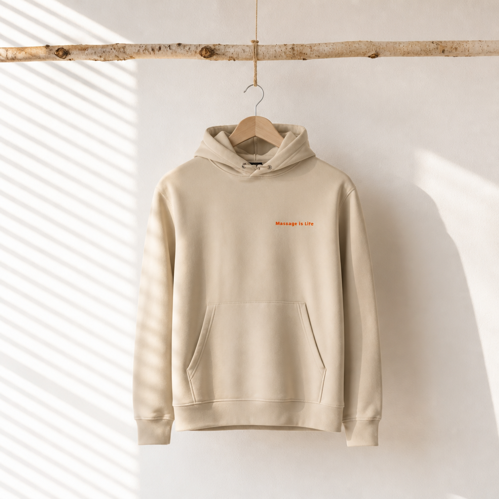 Sweat à capuche bio beige avec inscription 'Massage is Life', suspendu à une branche en bois, lumière naturelle