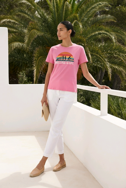 T-shirt rose bio Massage Is Life avec imprimé coucher de soleil, porté avec pantalon blanc et espadrilles, fond palmiers